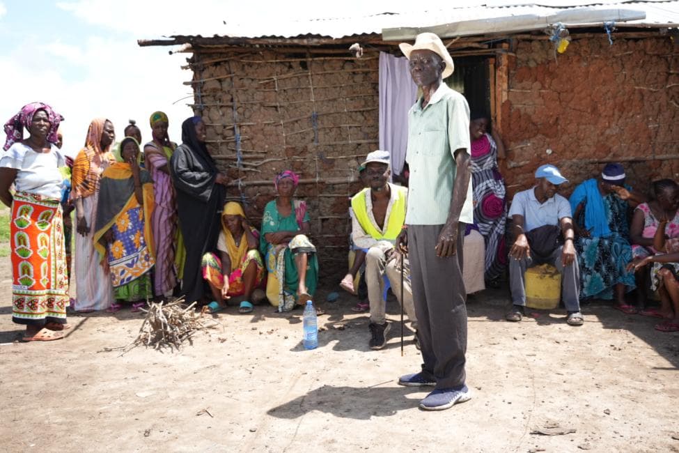 Photograph: women and men of a dryland community in Kenya gather outside a mud-walled house as an elder in a hat stands to address them.